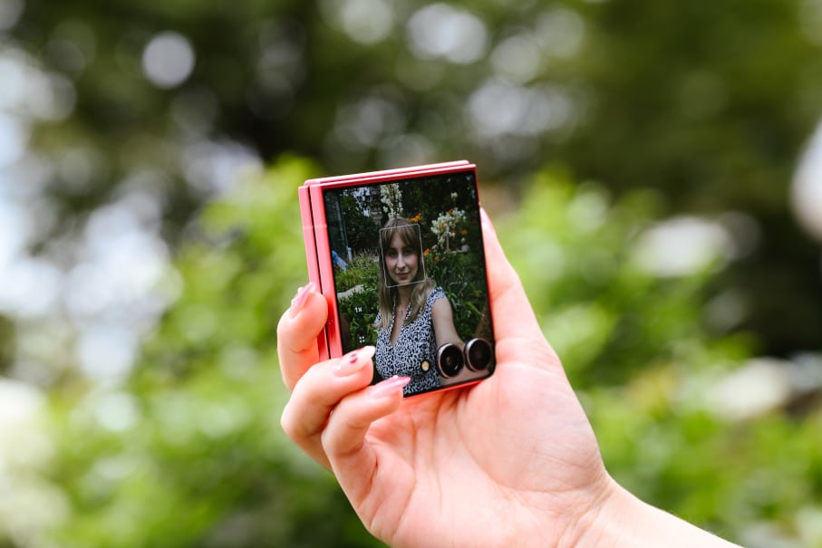 Ręka trzymająca różowy, składany smartfon z zewnętrznym ekranem przedstawiającym kobietę pozującą do selfie na zewnątrz, otoczoną rozmytymi zielonymi liśćmi.